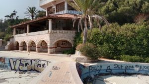Deserted villa with graffiti and empty pool, surrounded by greenery under a clear sky.