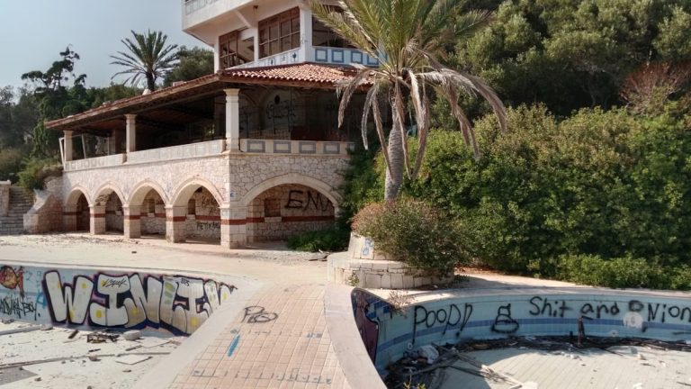 Deserted villa with graffiti and empty pool, surrounded by greenery under a clear sky.